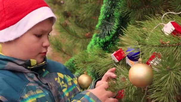 Um menino close-up em um chapéu de Papai Noel pendura uma bola festiva de prata. — Vídeo de Stock