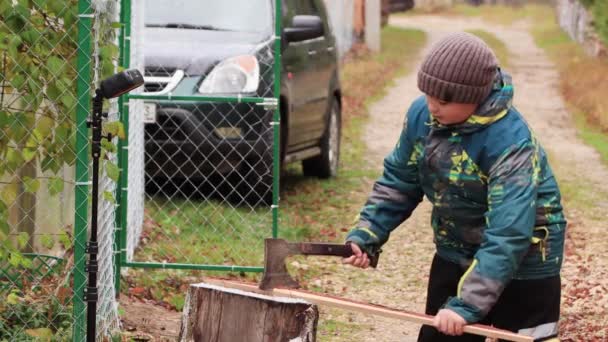 Een jongen op een boomstronk hakt onhandig een stok met een bijl. — Stockvideo