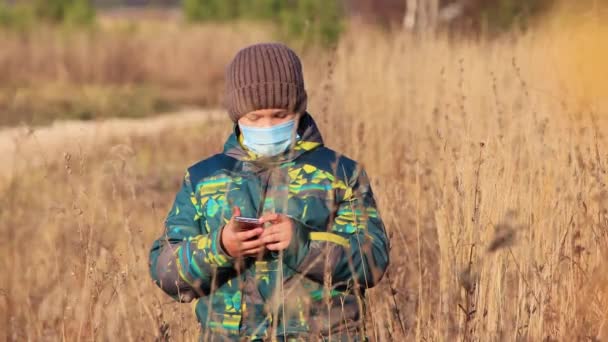 Ένα αγόρι σε μια ιατρική μάσκα, κρατώντας ένα τηλέφωνο στο πεδίο, κοιτάζοντας την κάμερα. — Αρχείο Βίντεο