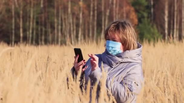 Una joven con una máscara, en la hierba alta, mira el teléfono. — Vídeos de Stock