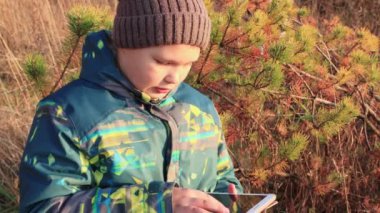 Elinde tabletle kırmızı-yeşil bir bitkinin arka planında duran bir çocuk..