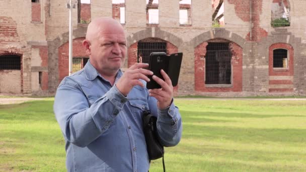 A man in a denim shirt looks at the phone against a destroyed red brick wall. — Stock Video