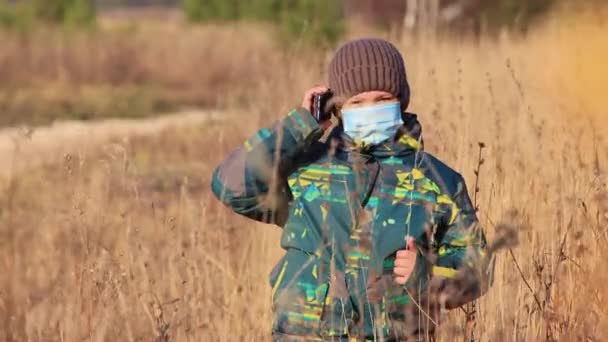 Un niño con una máscara médica la hierba alta, que está hablando por teléfono. — Vídeo de stock
