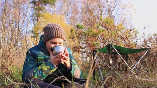 Chłopiec w kapeluszu i kurtce siedzi na trawie i pije herbatę z kubka. — Wideo stockowe
