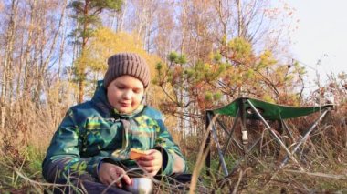 Şapkalı ve ceketli bir çocuk çimenlerde oturuyor, bir kupa takıyor ve kurabiye yiyor..