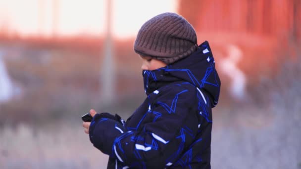Портрет щасливого хлопчика в куртці і капелюсі з телефоном в профілі . — стокове відео