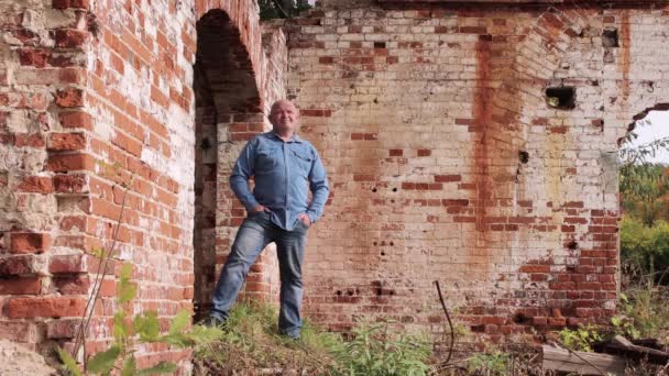 Um homem careca de jeans está na abertura de uma parede de tijolo vermelho arruinada — Vídeo de Stock