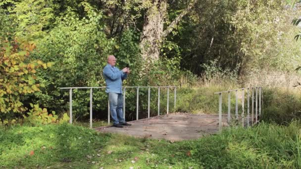 Muž v džínách stojí na mostě a mluví online na smartphonu. — Stock video