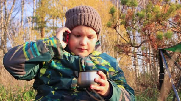 Chlapec v klobouku mluví po telefonu a pije z hrnku. — Stock video