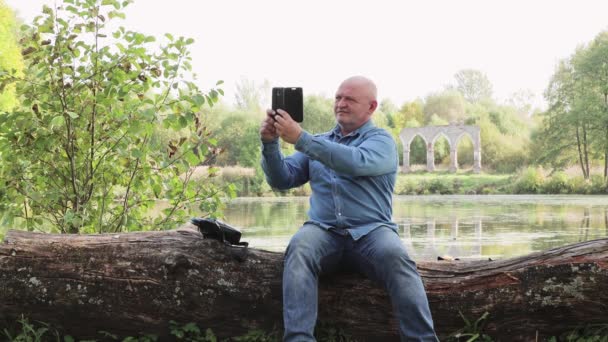 Muž s telefonem v ruce sedí na kládě u jezera.. — Stock video