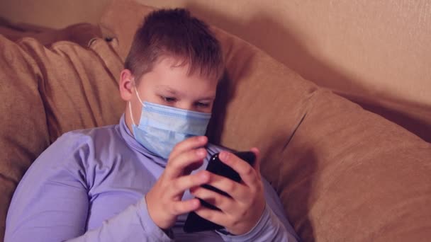 Barn dreng i en maske ved hjælp af en smartphone gennemser sociale netværk. – Stock-video
