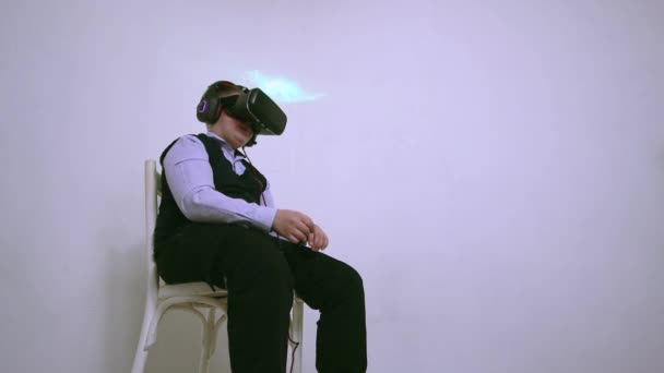 Een jongen in een virtual reality headset en koptelefoon speelt terwijl hij op een stoel zit — Stockvideo