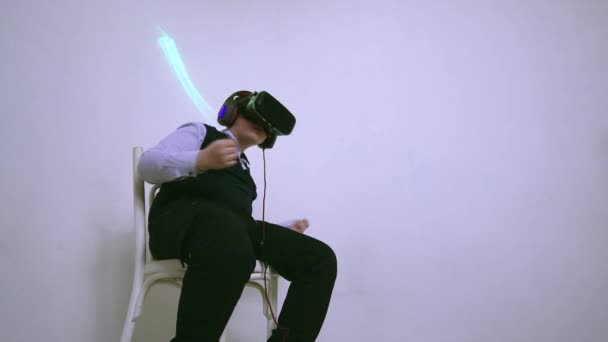 Een jongen in een virtual reality headset en een koptelefoon zittend op een stoel danst — Stockvideo