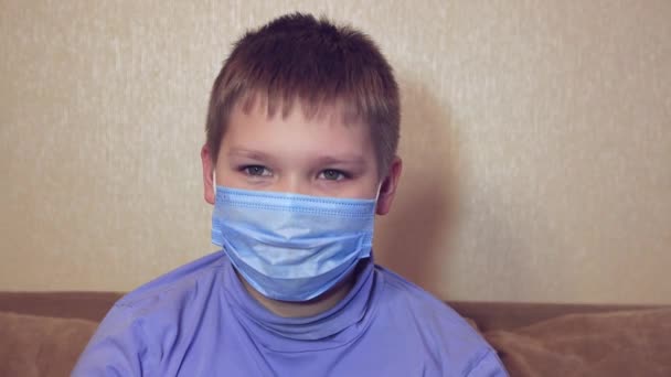 戴着医疗面具的快乐而轻松的男孩坐在沙发上笑着, — 图库视频影像