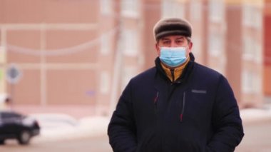 Tıbbi maske takmış bir adamın yakın plan fotoğrafı. Duruyor, ileri ve geri bakıyor..