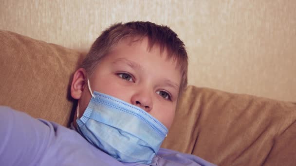 Egy boldog fiú orvosi maszkban fekszik a kanapén, és a kamerába néz.. — Stock videók