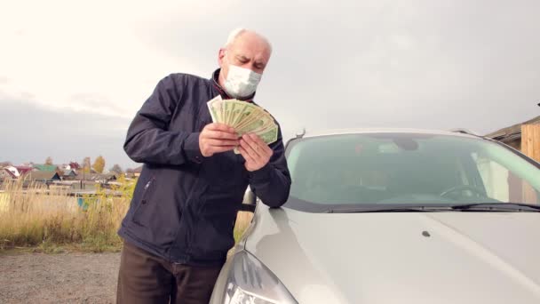 Ein glücklicher Mann mit Maske, an ein Auto gelehnt, zählt Dollar - sein Einkommen. — Stockvideo