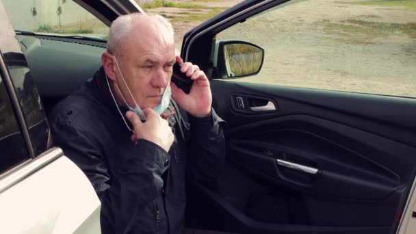 Podnikatel v masce, který sedí na prahu svého auta a mluví do telefonu. — Stock video
