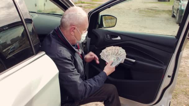 Бизнесмен в маске, сидящий на пороге машины, считает доллары. — стоковое видео