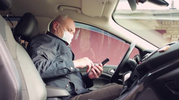 Un homme d'affaires avec un masque médical, conduisant une voiture, travaille en ligne sur un smartphone. — Video