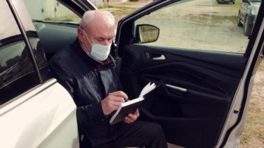Maskeli bir iş adamı arabanın kapısında oturmuş bir defter dolduruyor.