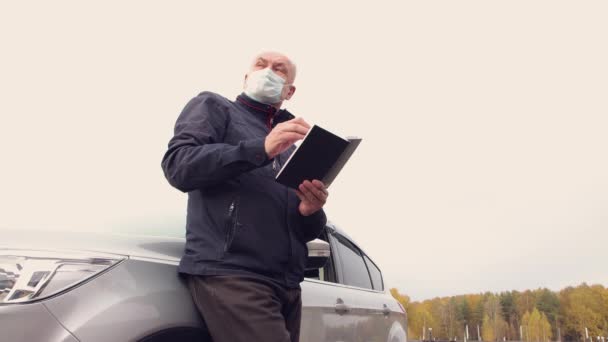 Ένας επιχειρηματίας με μάσκα, στέκεται δίπλα στο αυτοκίνητο, ξεφυλλίζοντας ένα σημειωματάριο.. — Αρχείο Βίντεο