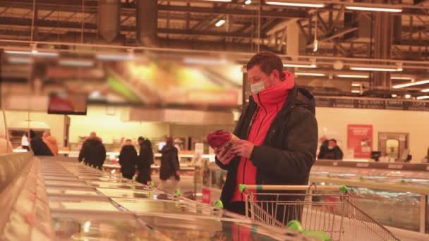 Un homme avec un chariot dans un supermarché lit l'emballage, avec un masque sur le visage. — Video