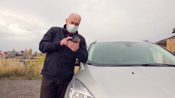 Business man in a medical mask standing by the car works online on a smartphone. — Stock Video