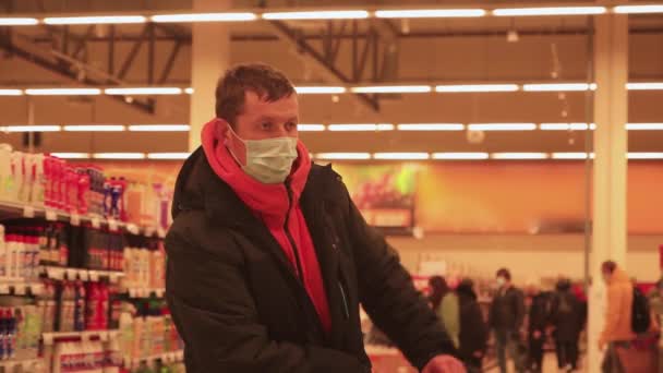Молодой человек стоит с тележкой в супермаркете, с маской на лице. — стоковое видео