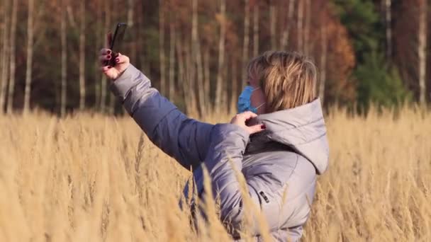 Kobieta w masce na twarzy robi sobie selfie w wysokiej trawie. — Wideo stockowe