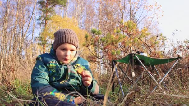 Een jongen zit in hoog gras tegen een achtergrond van geel gebladerte en eet. — Stockvideo