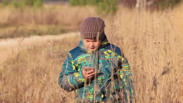 El chico en la hierba alta habla en línea por teléfono, señala con el dedo a su cabeza — Vídeo de stock