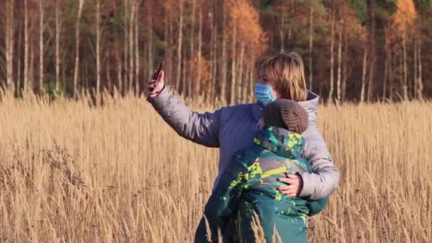 Moeder en zoon met maskers op hun gezicht in het veld nemen een selfie op een smartphone — Stockvideo