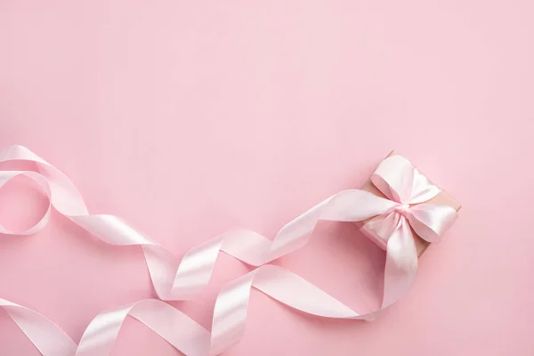 Pastel pembe arka planda uzun kurdeleli hediye kutusu. Sevgililer Günü, Anneler Günü ya da Kadınlar Günü hediyesi. Şenlikli tek renkli arka plan. Üst görünüm düz, boş alanı var..