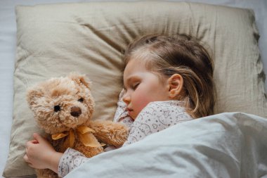 Yatakta uyuyan küçük kız evde yumuşak oyuncakları kucaklıyor.