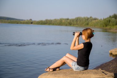 bir taş üzerine oturan genç kız nehir yakınında ve dürbün ile görünüyor
