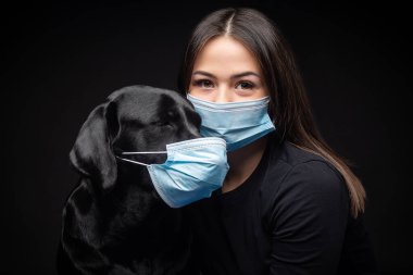Labrador Retriever köpeğinin portresi. Koruyucu bir tıbbi maske takmış, bir de kadın sahibi var. Fotoğraf stüdyosunda çekilmişti.