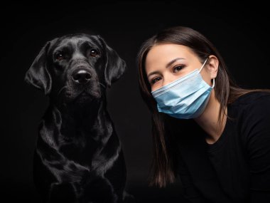 Labrador Retriever köpeğinin portresi. Koruyucu bir tıbbi maske takmış, bir de kadın sahibi var. Fotoğraf stüdyosunda çekilmişti.