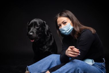 Labrador Retriever köpeğinin portresi. Koruyucu bir tıbbi maske takmış, bir de kadın sahibi var. Fotoğraf stüdyosunda çekilmişti.