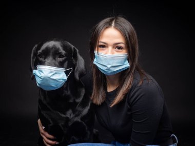 Labrador Retriever köpeğinin portresi. Koruyucu bir tıbbi maske takmış, bir de kadın sahibi var. Fotoğraf stüdyosunda çekilmişti.