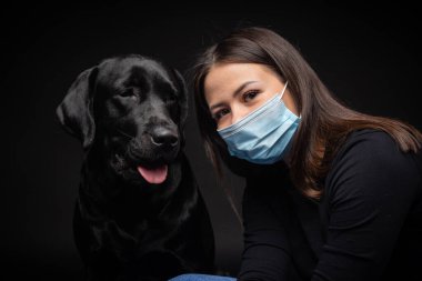 Labrador Retriever köpeğinin portresi. Koruyucu bir tıbbi maske takmış, bir de kadın sahibi var. Fotoğraf stüdyosunda çekilmişti.