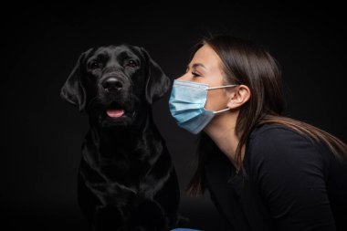 Labrador Retriever köpeğinin portresi. Koruyucu bir tıbbi maske takmış, bir de kadın sahibi var. Fotoğraf stüdyosunda çekilmişti.