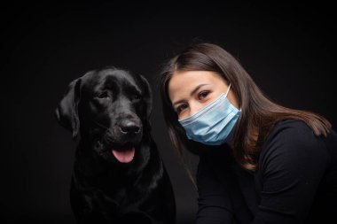 Labrador Retriever köpeğinin portresi. Koruyucu bir tıbbi maske takmış, bir de kadın sahibi var. Fotoğraf stüdyosunda çekilmişti.