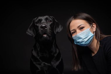 Labrador Retriever köpeğinin portresi. Koruyucu bir tıbbi maske takmış, bir de kadın sahibi var. Fotoğraf stüdyosunda çekilmişti.