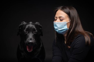 Labrador Retriever köpeğinin portresi. Koruyucu bir tıbbi maske takmış, bir de kadın sahibi var. Fotoğraf stüdyosunda çekilmişti.