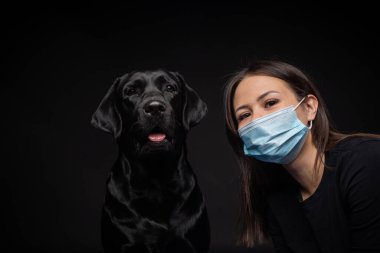 Labrador Retriever köpeğinin portresi. Koruyucu bir tıbbi maske takmış, bir de kadın sahibi var. Fotoğraf stüdyosunda çekilmişti.