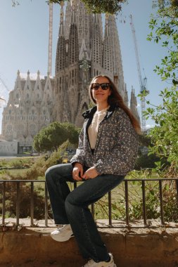 Barselona 'da Sagrada Familia' nın önündeki parmaklıklarda oturan genç bir kadın ve yeşil bir gölet.
