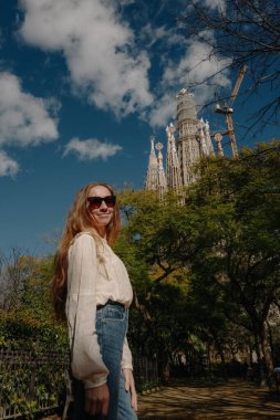 Parkta gülümseyen genç bir kadın boho bluz ve Sagrada Familia yakınlarında koyu renk kot pantolon giyiyor.