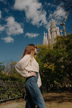 Kameraya arkası dönük genç bir kadın parkta Sagrada Familia 'ya bakıyor.