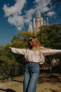 Parkta gülümseyen genç bir kadın boho bluz ve Sagrada Familia yakınlarında koyu renk kot pantolon giyiyor.